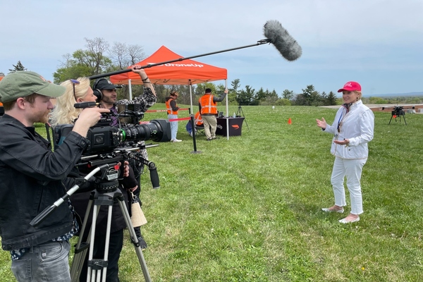 A woman stands in front of a camera crew. In the background, stands an orange tent sporting the words "DroneUp" next to a drone that appears to be preparing to take off.